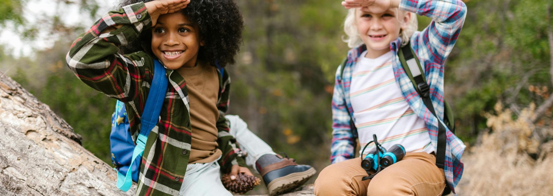 Two children sit on a tree trunk in a forest, engaged in a playful outdoor adventure.