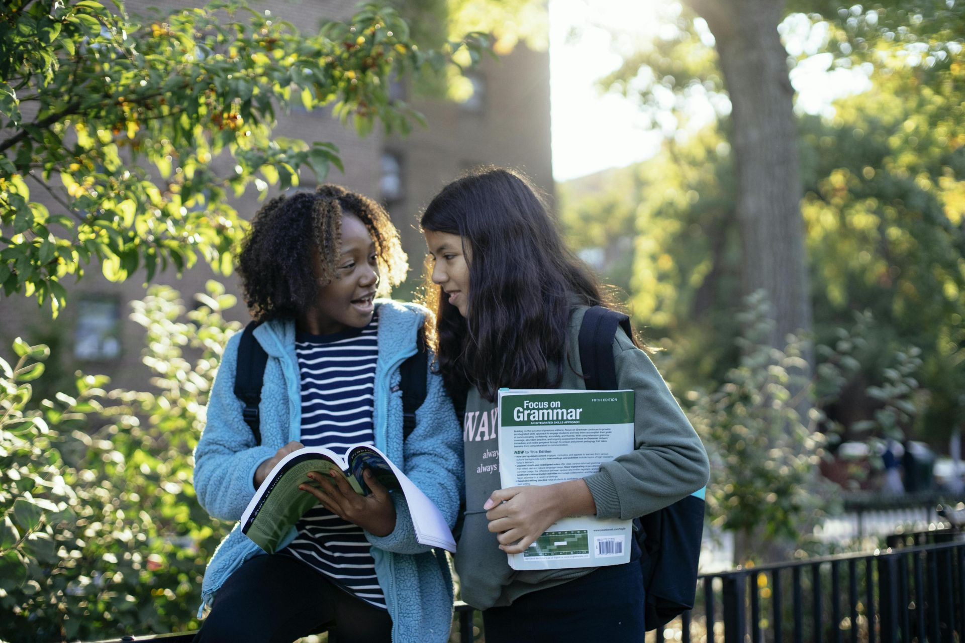 Cheerful multiracial friends in casual clothes with backpacks standing near fence in park while talking about study